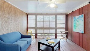 Una habitación con un sofá azul, una mesita con libros, un ventilador de techo y un ventanal con persianas.