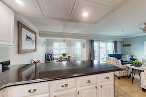 A modern kitchen with white cabinetry, a dark countertop, and a dining area with a blue sofa and patterned armchair. - White Marlin 406B (Ocean City)