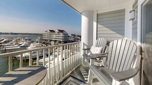 A balcony with two Adirondack chairs, a view of a marina with boats, and a building with a red roof in the background. - Luxury - bay view condo with balcony, pool, W/D, AC & a short walk to the beach (Ocean City)