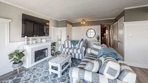 A living room with a fireplace, striped sofa, and a television mounted on the wall. - Luxury - bay view condo with balcony, pool, W/D, AC & a short walk to the beach (Ocean City)