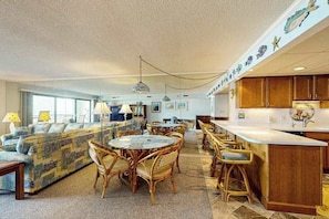 A spacious living area with a sofa, dining table, and chairs, a kitchen with wooden cabinets, and a decorative ceiling. - Spacious, oceanfront condo with balcony, AC, full kitchen & stunning beach views (Ocean City)