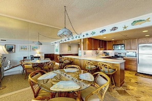 A kitchen with wooden cabinets, a central island, and a dining area with wicker chairs and a glass table. - Spacious, oceanfront condo with balcony, AC, full kitchen & stunning beach views (Ocean City)
