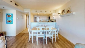 A dining area with a table and chairs, a kitchen in the background, and a hallway leading to another room. - Triton's Trumpet B502 (Ocean City)