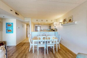 Uma área de refeições com mesa e cadeiras, uma cozinha ao fundo e um corredor que leva a outro quarto.