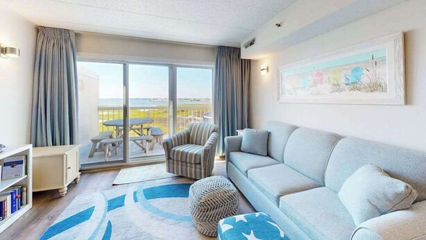 A living room with a sofa, ottoman, and striped armchair. There is a balcony with a table and chairs, and a bookshelf with books. - Triton's Trumpet B502 (Ocean City)
