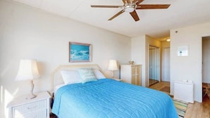 A bedroom with a bed, nightstands, a ceiling fan, a painting, and a door leading to another room. - Triton's Trumpet B502 (Ocean City)