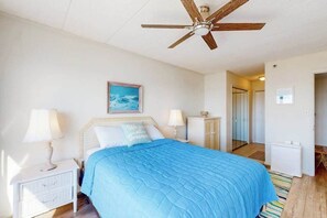 Quarto com cama, mesas de cabeceira, ventilador de teto, um quadro e uma porta que dá para outro cômodo.