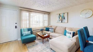 A living room with a sofa, a coffee table, and a blue armchair. There is a clock on the wall and a window with blinds. - Bonnie Belle 208 (Ocean City)