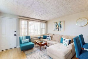 Ein Wohnzimmer mit einer Couch, einem Couchtisch und einem blauen Sessel. An der Wand hängt eine Uhr und es gibt ein Fenster mit Jalousien.