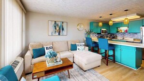 A modern living room with a sofa, ottoman, and coffee table, a kitchen with teal cabinets and bar stools, and a large window with blinds. - Bonnie Belle 208 (Ocean City)