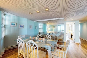 A spacious dining area with a glass table and white wicker chairs, a wooden ceiling, and light blue walls. - Beachfront Coastal Retreat w/Direct Beach Views, Shared Pool, Balcony, WiFi & AC (Ocean City)