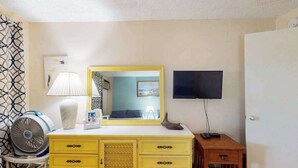 Um quarto com cômoda amarela, espelho, televisão, ventilador e porta.