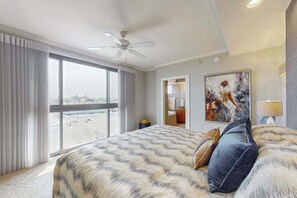 A hotel room with a large bed, a ceiling fan, a painting, and a view of the city through large windows. - Luxurious Ocean Front Condo w/Beach Access, Central AC & Gas Fireplace (Ocean City)