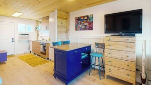 Una cocina compacta con una isla azul, una zona de comedor con sillas, un televisor y un cuadro en la pared.