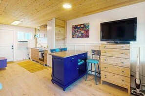 Eine kleine Küche mit einer blauen Kücheninsel, ein Essbereich mit Stühlen, ein Fernseher und ein Bild an der Wand.