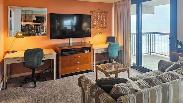 Een hotelkamer met een televisie, bureau, stoel, bank en een balkon met uitzicht.
