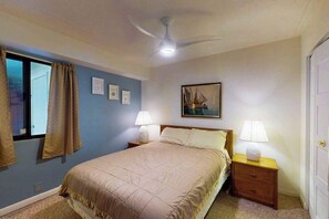 Ein Schlafzimmer mit einem Bett, zwei Nachttischen mit Lampen, einem Deckenventilator, einem Fenster mit Vorhängen und einem gerahmten Bild an der Wand.
