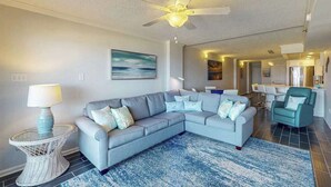 A living room with a sectional sofa, a round coffee table, a ceiling fan, and a kitchen area in the background. - Stunning Ocean front condo w/beach & ocean views, AC & shared pool (Ocean City)