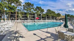 Une piscine entourée d’arbres et de bâtiments, avec des chaises longues et des parasols.