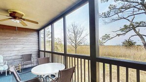 Un espace extérieur abrité, équipé d’une table et de chaises, d’un ventilateur de plafond et de grandes fenêtres offrant une vue sur les arbres et la pelouse.