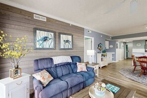 A living room with a blue sofa, white side tables, and a dining area with a wooden table and chairs. - Aventurra 206 (Ocean City)