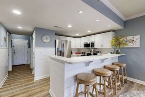 A modern kitchen with white cabinetry, a central island with stools, a stainless steel refrigerator, and a microwave. - Aventurra 206 (Ocean City)