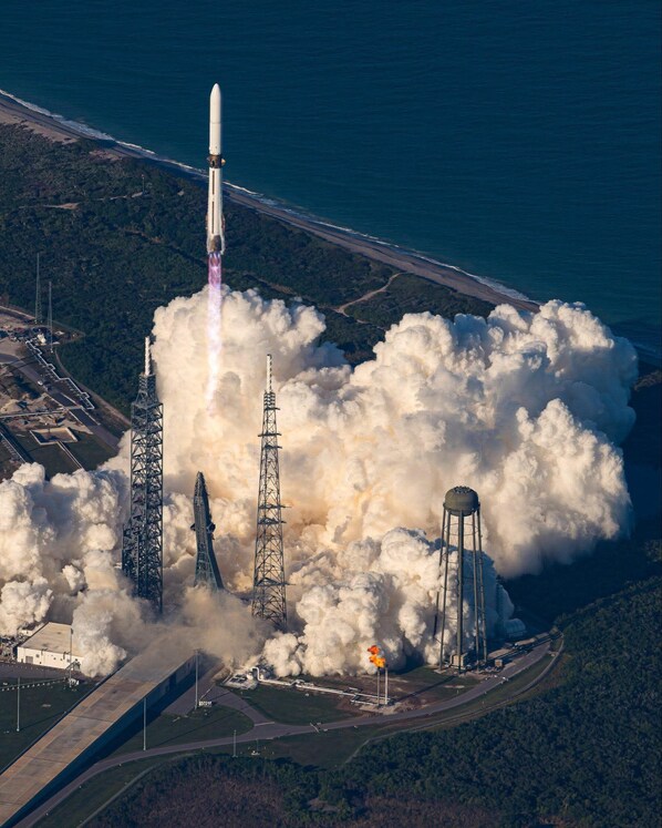 A rocket launch with a launch tower, smoke, and a body of water in the background.