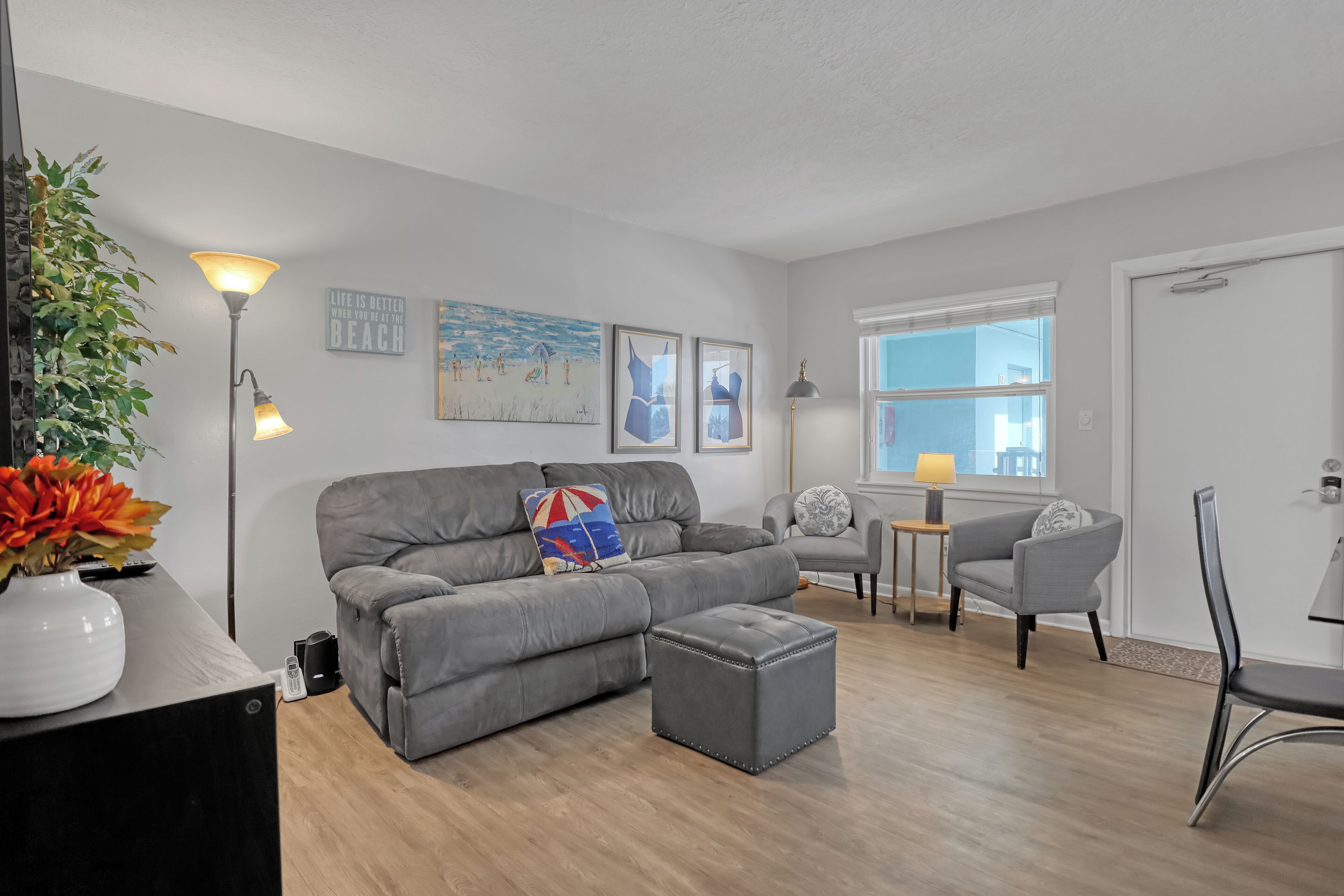 Ein modernes Wohnzimmer mit grauem Sofa, Hocker und Sesseln. Ein kleiner Tisch mit Lampe, einer Zimmerpflanze und Kunstwerken an den Wänden.