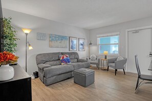 Un salon moderne avec un canapé gris, un pouf et des fauteuils. On y trouve une petite table avec une lampe, une plante en pot et des œuvres d’art accrochées aux murs.