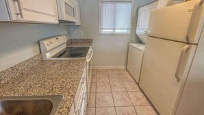 A compact kitchen with white appliances, a granite countertop, and a window with blinds. - Blaire Isle 302N (Ocean City)