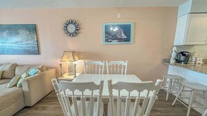 A living room with a beige sofa, a dining table with white chairs, a wall clock, and a framed picture. - Blaire Isle 302N (Ocean City)