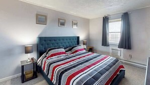 Un dormitorio con cabecera capitonada, colcha a rayas, lámparas de noche, una mesita y una ventana con cortinas.