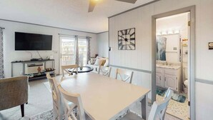 Un comedor con una mesa y sillas, un ventilador de techo, un televisor de pantalla plana y vistas al exterior a través de un ventanal.