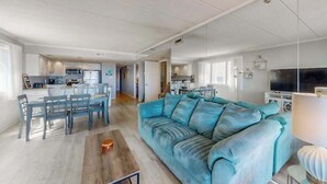 Amplia sala de estar con sofá, mesa de comedor y sillas, una cocina al fondo y un televisor a la derecha.