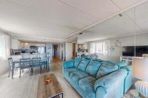 A spacious living area with a sofa, dining table, and chairs, a kitchen in the background, and a TV on the right. - Oceanfront Condo with Balcony, Beach Access, Central AC, WiFi & Washer/Dryer (Ocean City)