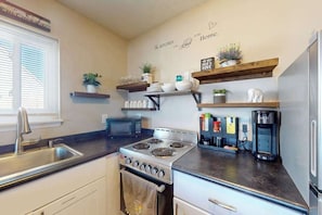 A kitchen with a stove, sink, microwave, and shelves with plants and kitchenware. - Sunny - 3rd floor, spacious condo with kitchen, balcony & canal views (Ocean City)