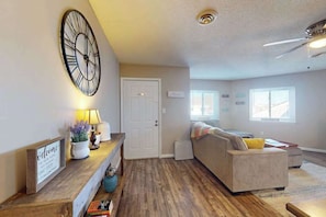 A living room with a clock, a sofa, a coffee table, and a door. - Sunny - 3rd floor, spacious condo with kitchen, balcony & canal views (Ocean City)