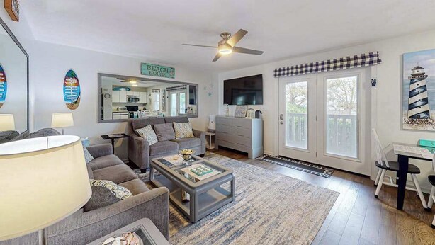 A living room with a sofa, coffee table, and a TV. There's a sign that says 'Beach House' and a window with a view. - Marathon North 103 (Ocean City)