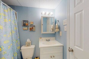 A bathroom with a toilet, sink, and mirror. There are wall decorations and a shower curtain with a fish pattern. - Harbor Sails 108 (Ocean City)