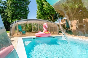 Una piscina con cubierta transparente, un flotador de flamenco rosa y un recinto de cristal con una silla.