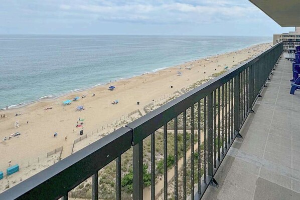 Blick auf einen Strand von einem Balkon mit Geländer, Strandstühlen und einem wolkenlosen Himmel.