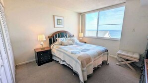 Une chambre à coucher avec un lit, deux tables de chevet avec des lampes, une fenêtre donnant sur la mer et un petit banc.
