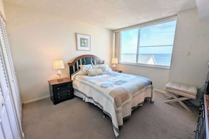 A bedroom with a bed, two bedside tables with lamps, a window with a view of the sea, and a small bench.