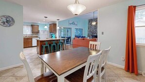 A dining area with a wooden table and white chairs, a kitchen in the background, and a living room with a sofa and a fireplace. - Teal Haven (Ocean Pines)