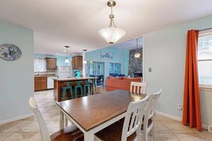 A dining area with a wooden table and white chairs, a kitchen in the background, and a living room with a sofa and a fireplace.