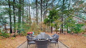 An outdoor patio with a table and chairs, surrounded by trees and a house in the background. - Teal Haven (Ocean Pines)