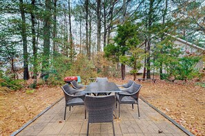 Une terrasse extérieure avec une table et des chaises, entourée d’arbres et avec une maison en arrière-plan.