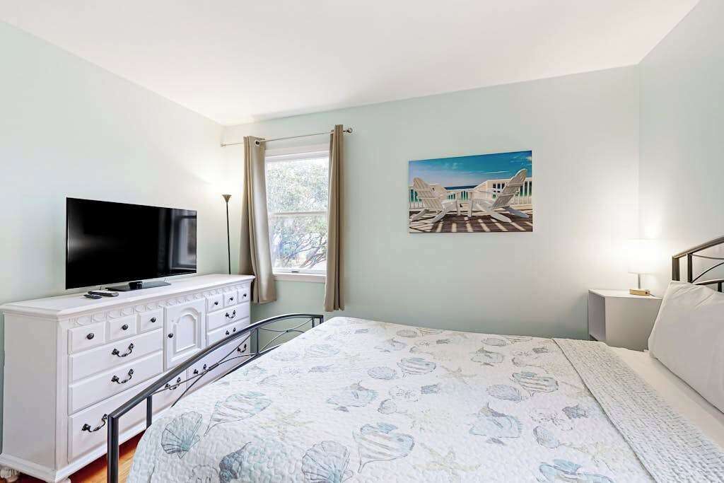 A bedroom with a bed, a dresser, a TV, a window with curtains, and a wall-mounted picture.