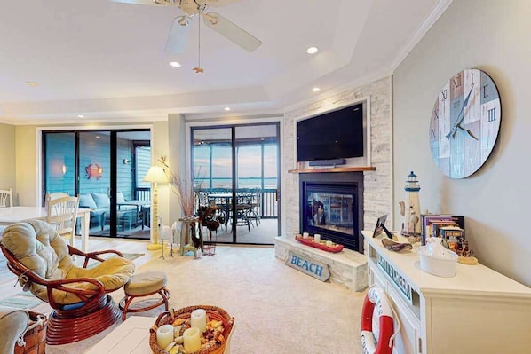 A spacious living room with a fireplace, a large clock, and a view through the glass doors. - Coastal condo with breathtaking views, balcony, fireplace, & W/D - pool access (Ocean City)