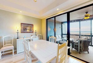 A dining area with a white table and chairs, a framed picture on the wall, and a view of the ocean through a glass door. - Coastal condo with breathtaking views, balcony, fireplace, & W/D - pool access (Ocean City)